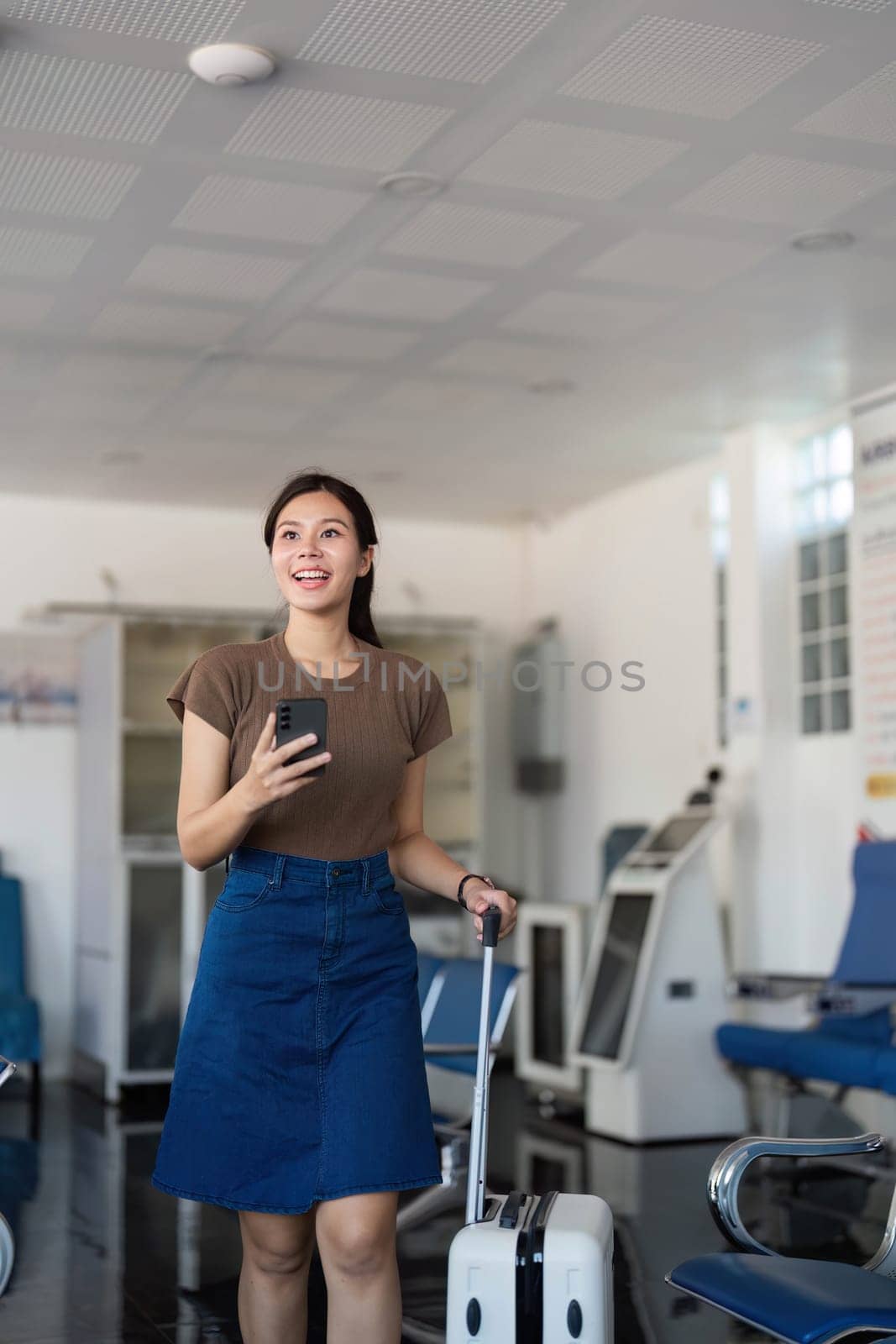 A cheerful young woman walking through the airport with her suitcase, checking her phone as she approaches the terminal gate.