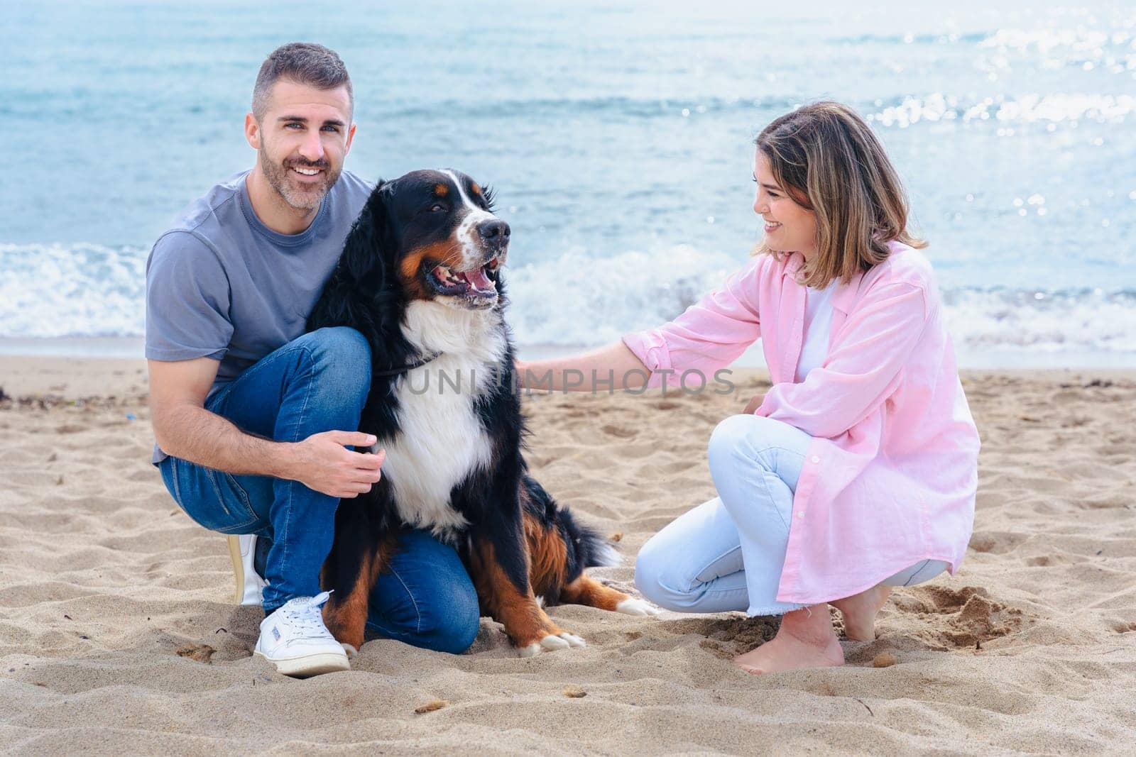 Portrait of a smiling caucasian couple with their large dog on the beach during vacation. Relaxed and joyful moment by the sea, with all three looking at the camera on a bright day.