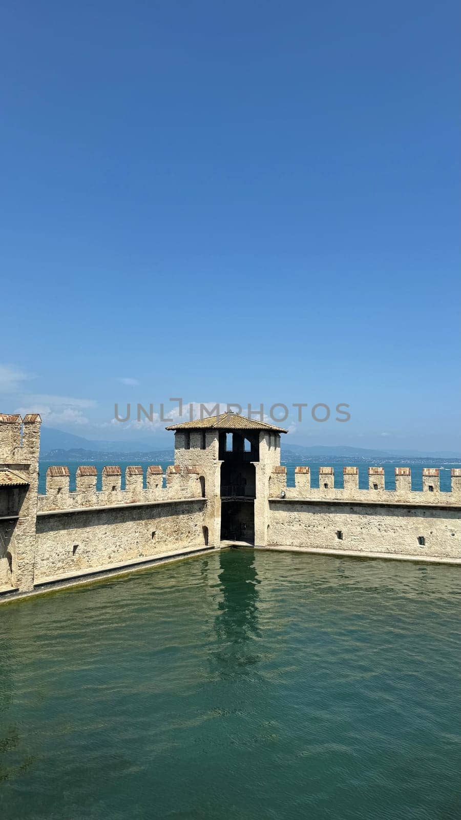 02.08.2024 Italy: Medieval Italian Castle Scaligero Walls Overlooking a Calm Blue Lake Garda Under a Clear Sky. A picturesque view of ancient 13th Century castle walls and tower surrounded by water, with scenic blue sky and serene lake Garda landscape in Sirmione, evoking a sense of history, tranquility, and natural beauty.