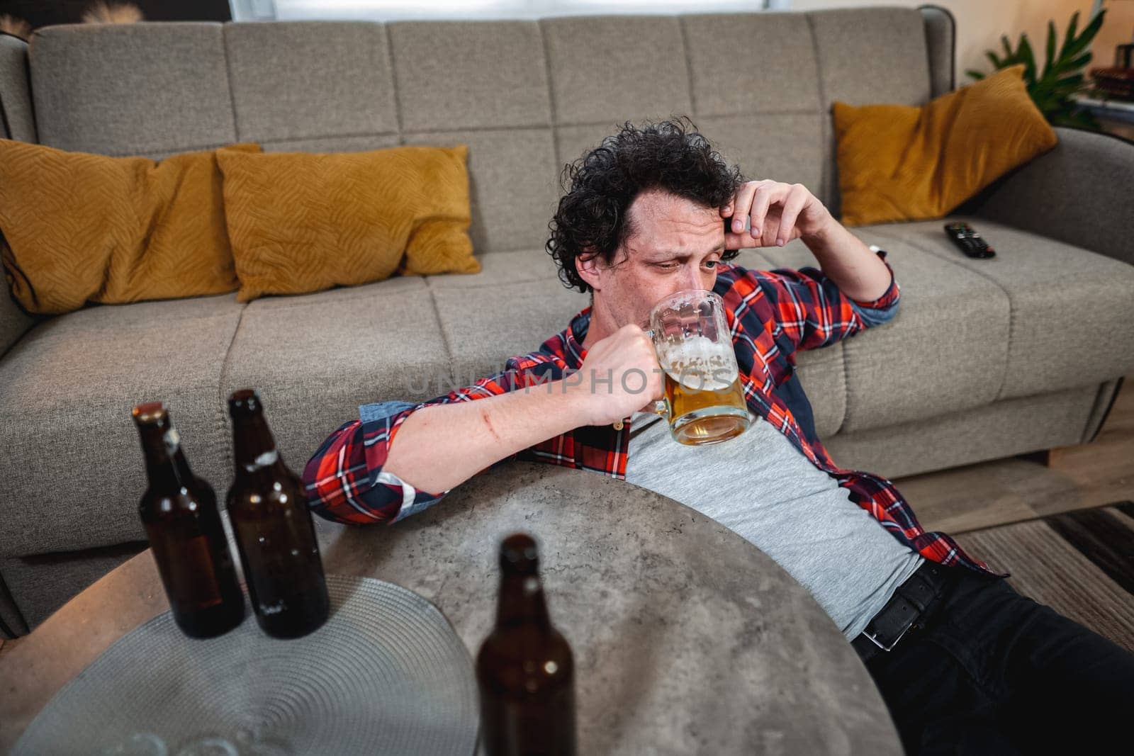 Young man drinking beer at home after party, lying on the floor by the sofa