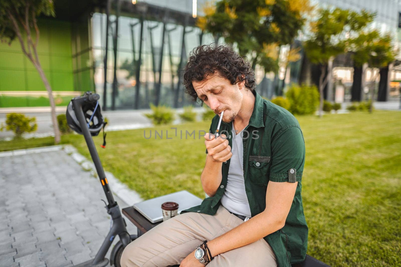 Man with curly hair sits on a park bench, lights a cigar and rests after riding an electric scooter