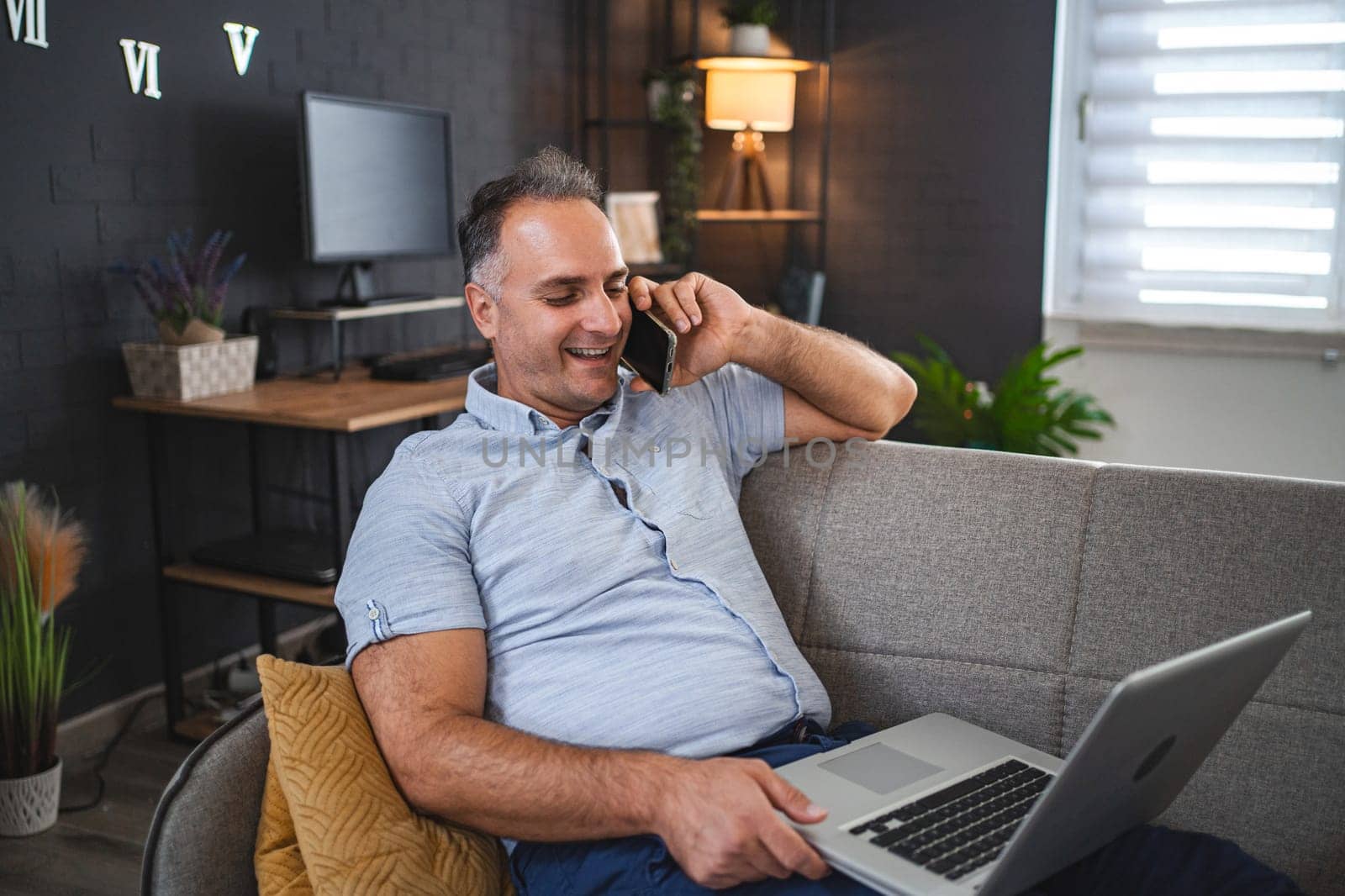 Middle aged Caucasian man talking on the phone while using laptop on the sofa.