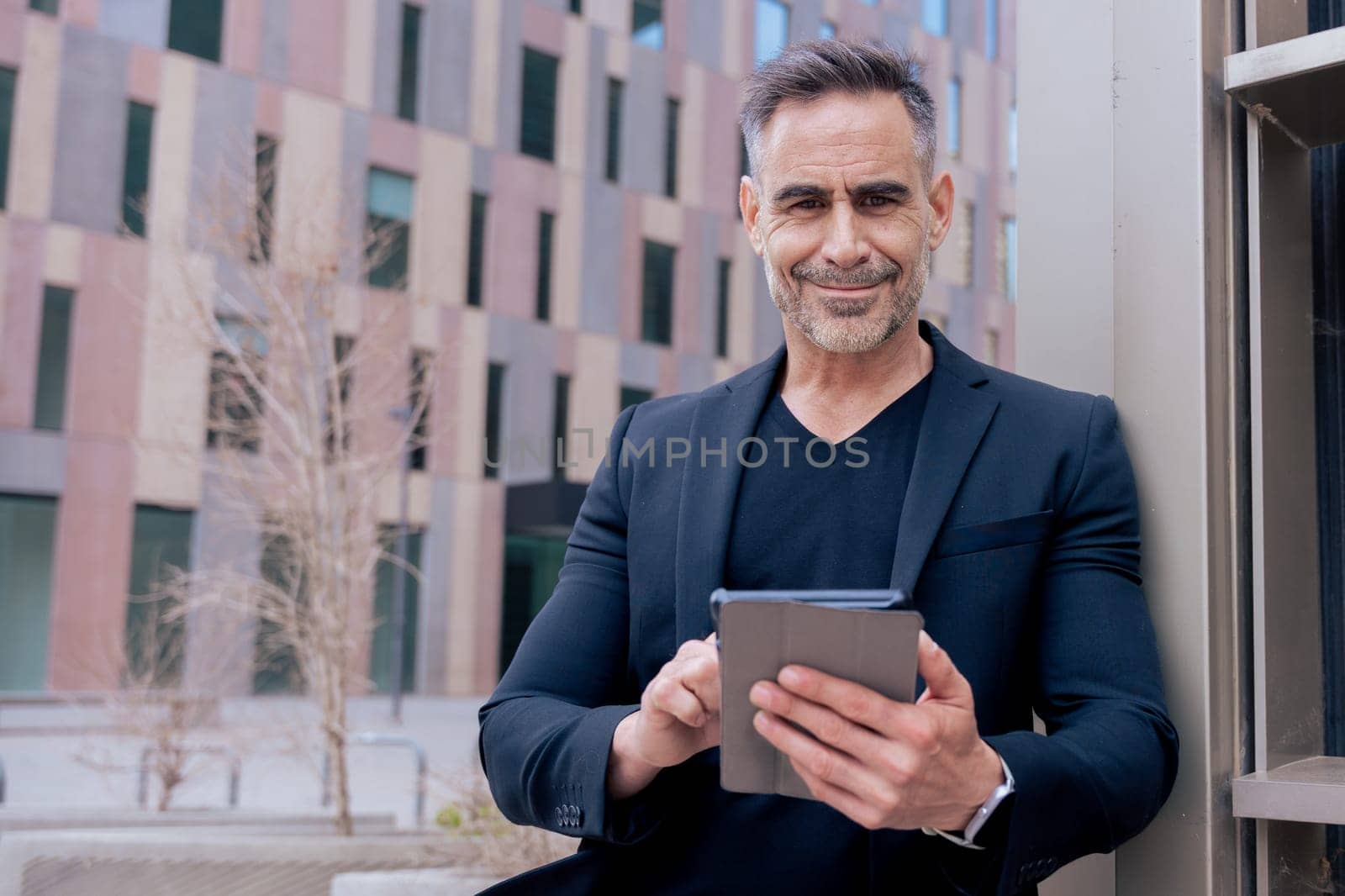 Confident businessman smiling while using a tablet outdoors in a modern urban setting.He wears a black suit and smartwatch. Concept of success, technology,digital lifestyle,and productivity.Copy space
