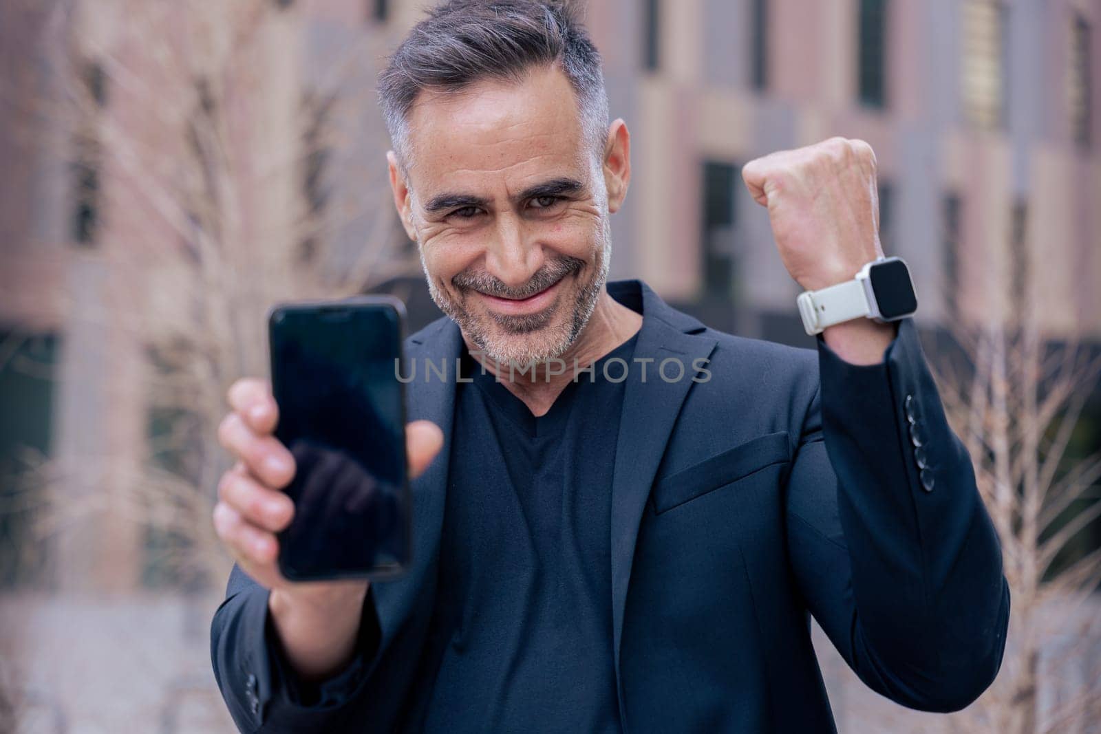 Confident businessman celebrates winning an online bet, holding a smartphone and raising his fist. He smiles outdoors in a city setting. Concept of success, digital lifestyle, and modern finance.
