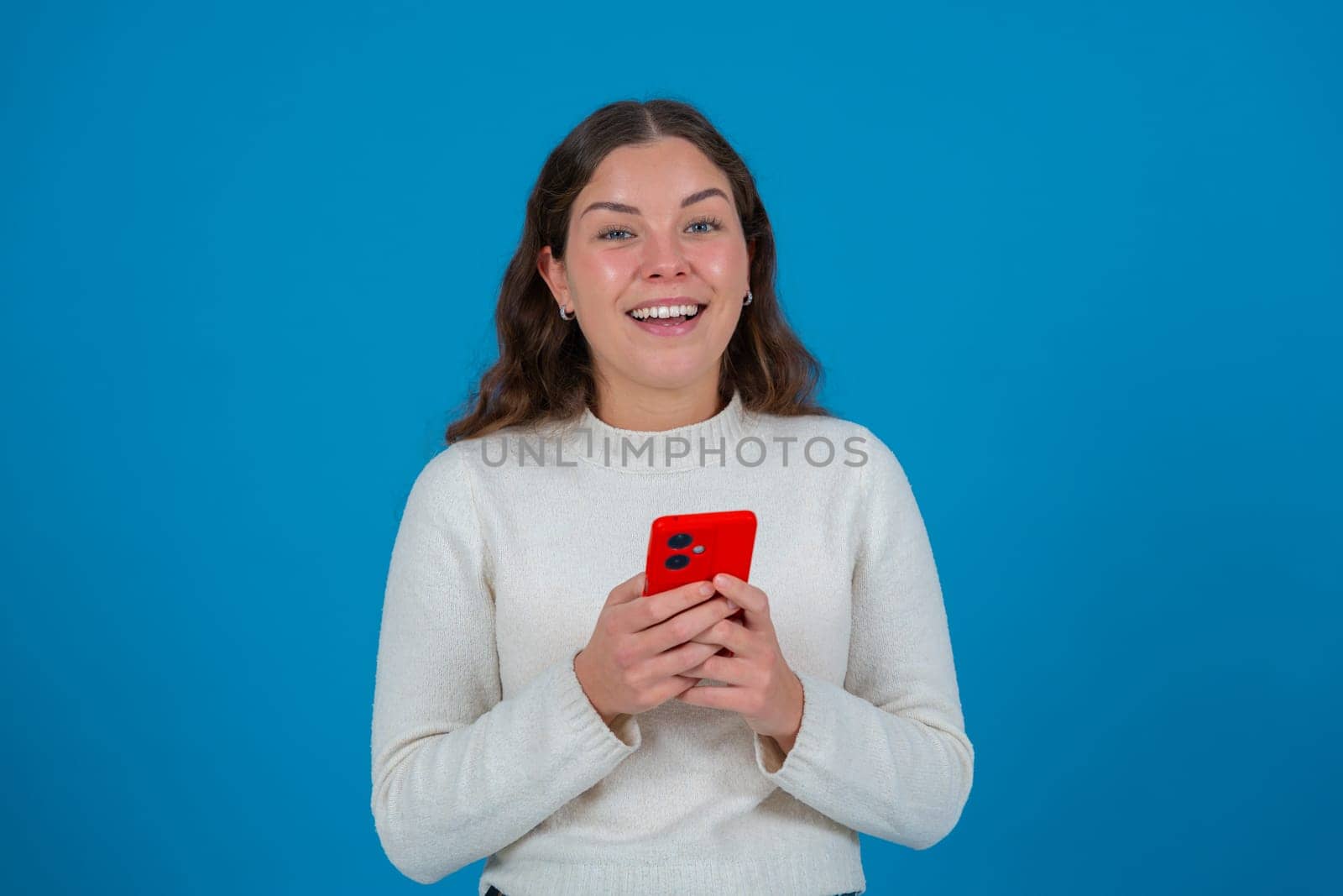 Cheerful young woman in white sweater looking at camera while holding a red smartphone in her hands, standing against a vivid blue background, representing connection, joy, and modern communication.