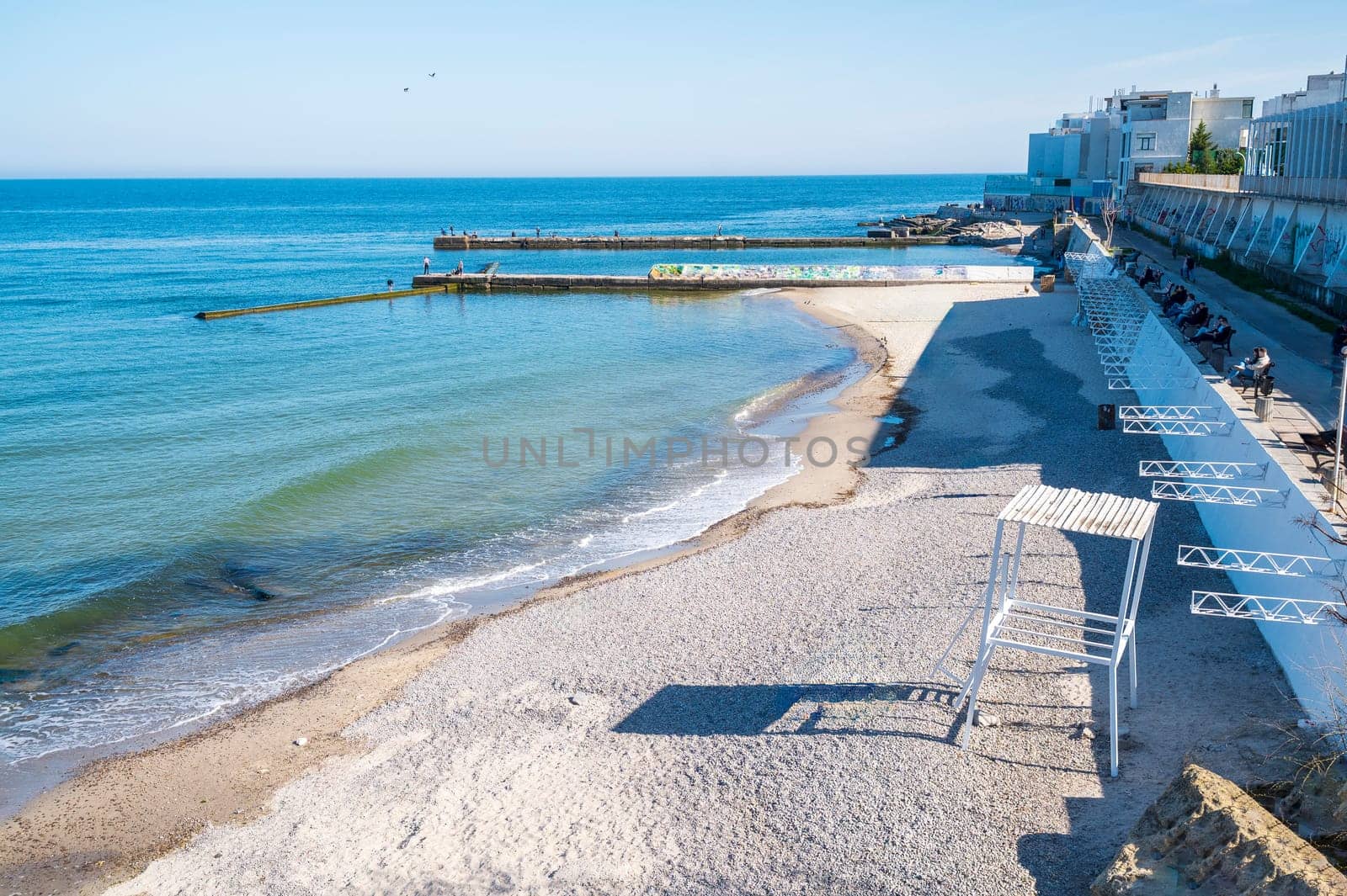Embankment, sea sand beach in European city. People are walking in the distance. Sandy beach in midday sun. Concrete jetties go into calm sea.