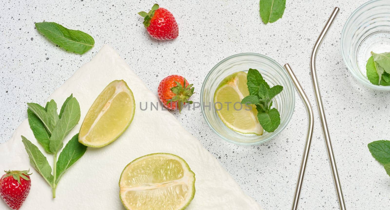 A top down view of ingredients for a refreshing drink, including whole and sliced limes, fresh mint sprigs, two empty glasses, and two metal straws