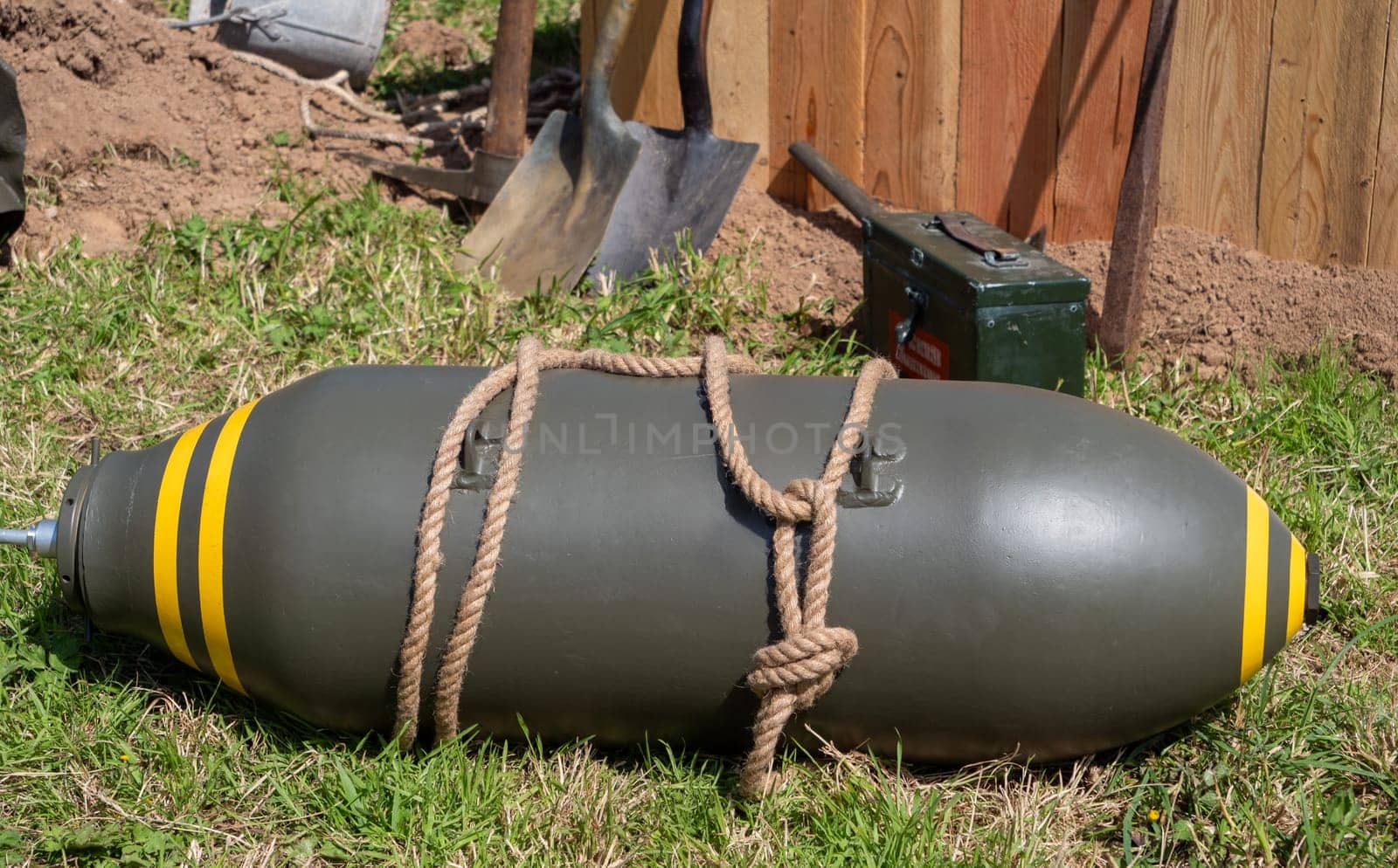 A large bomb is tied to a rope and is laying on the grass. The bomb is surrounded by a pile of dirt and a shovel. The scene is eerie and unsettling, as it suggests a potential danger or threat