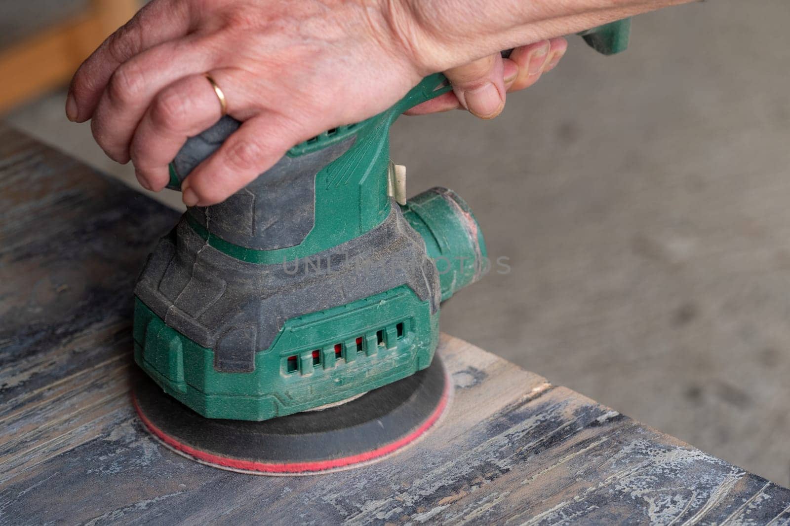 A man is using a sander to smooth out a surface. The sander is green and has a red handle. The man is wearing a ring on his finger