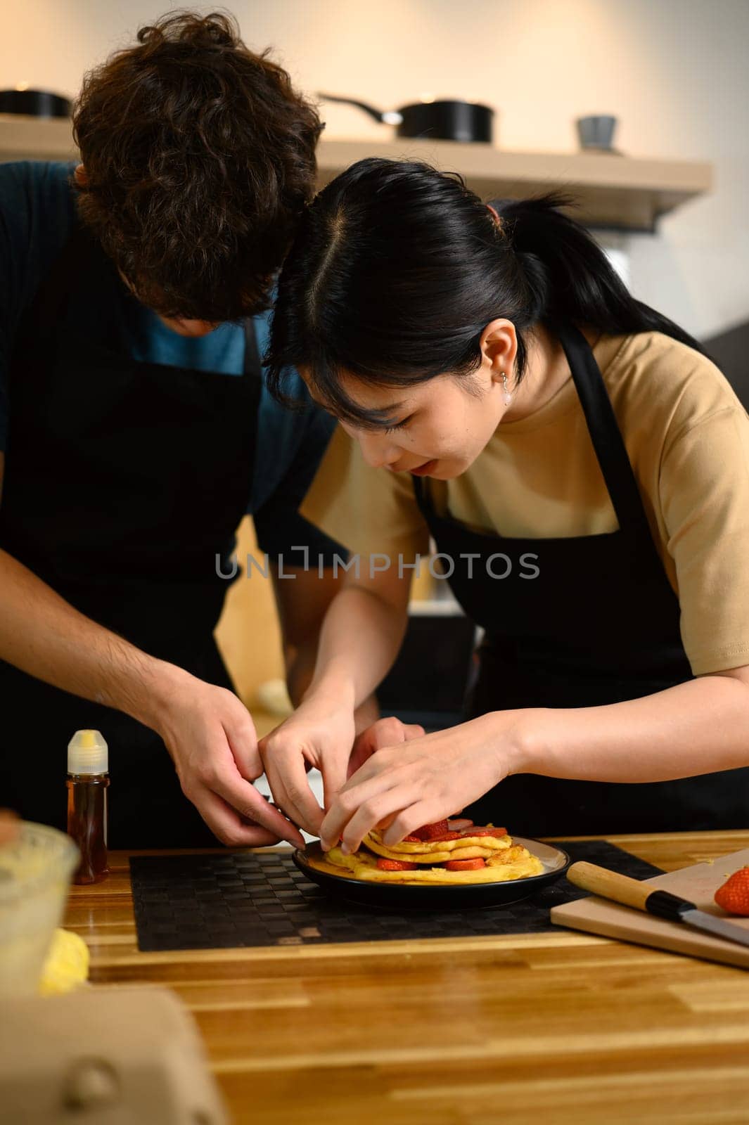 Loving young couple decorating homemade pancakes with sliced strawberries together.