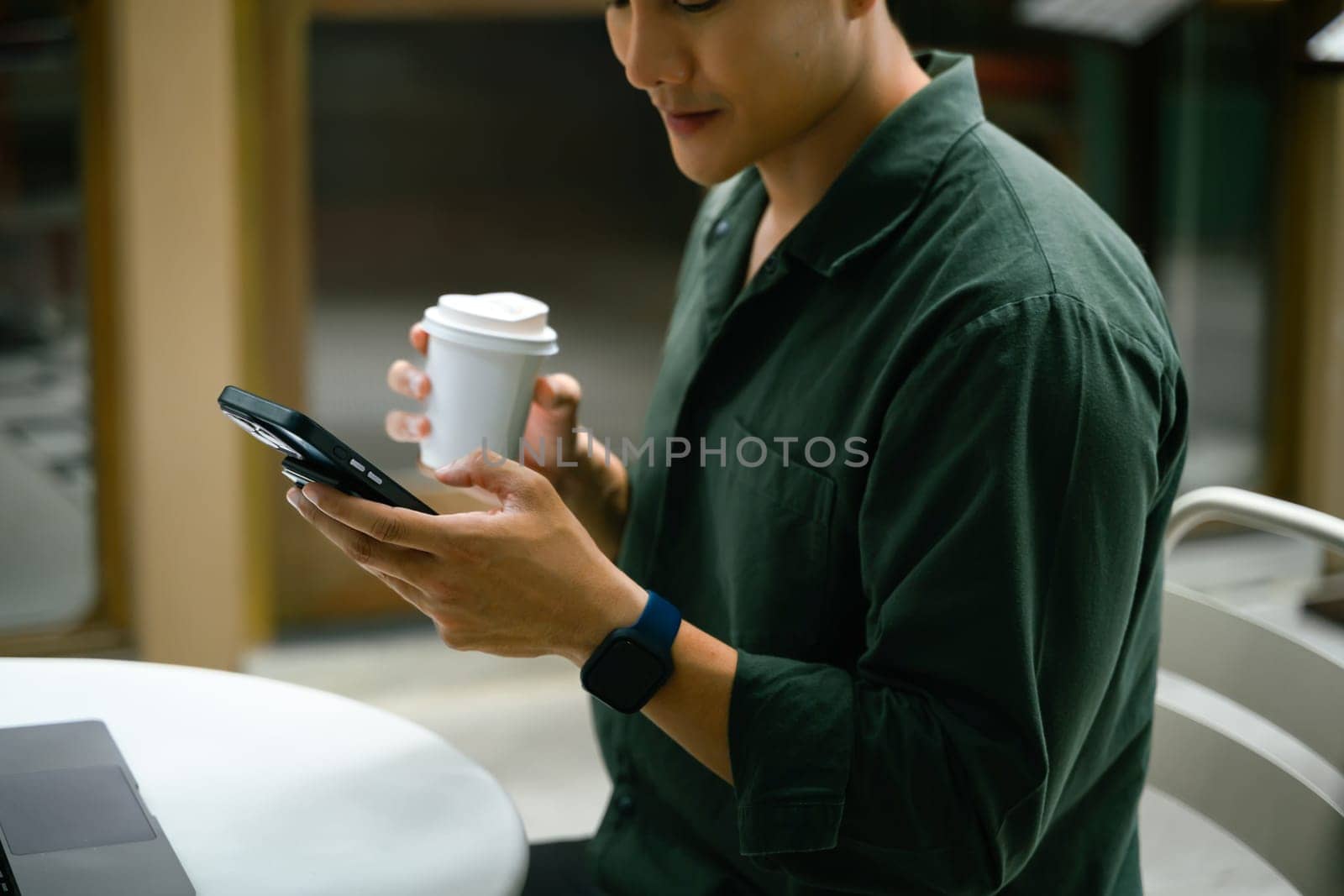 Man holding a smartphone and takeaway coffee cup while sitting at an outdoor cafe.