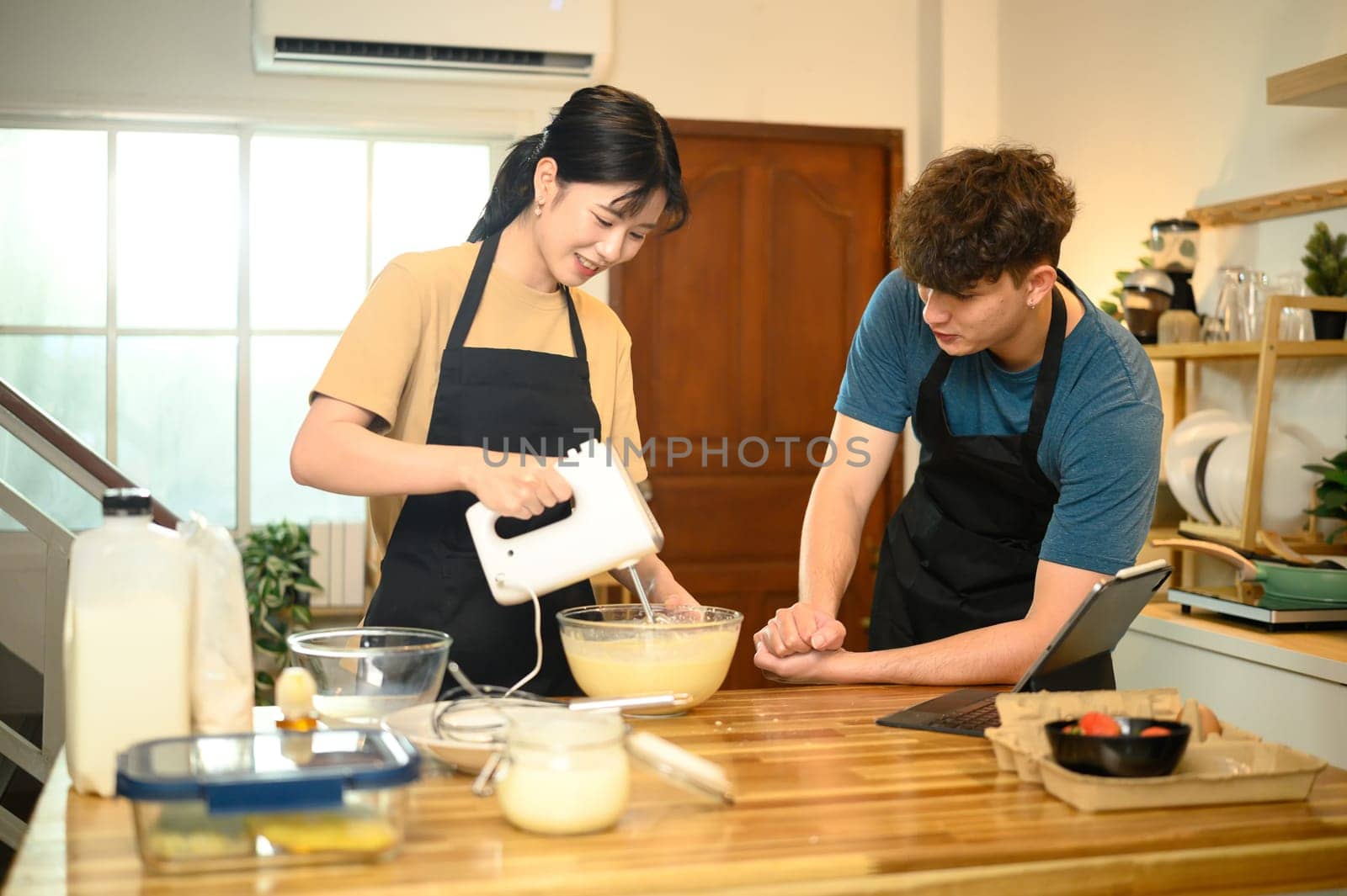 Smiling couple making pancake batter together using a hand mixer in a kitchen.