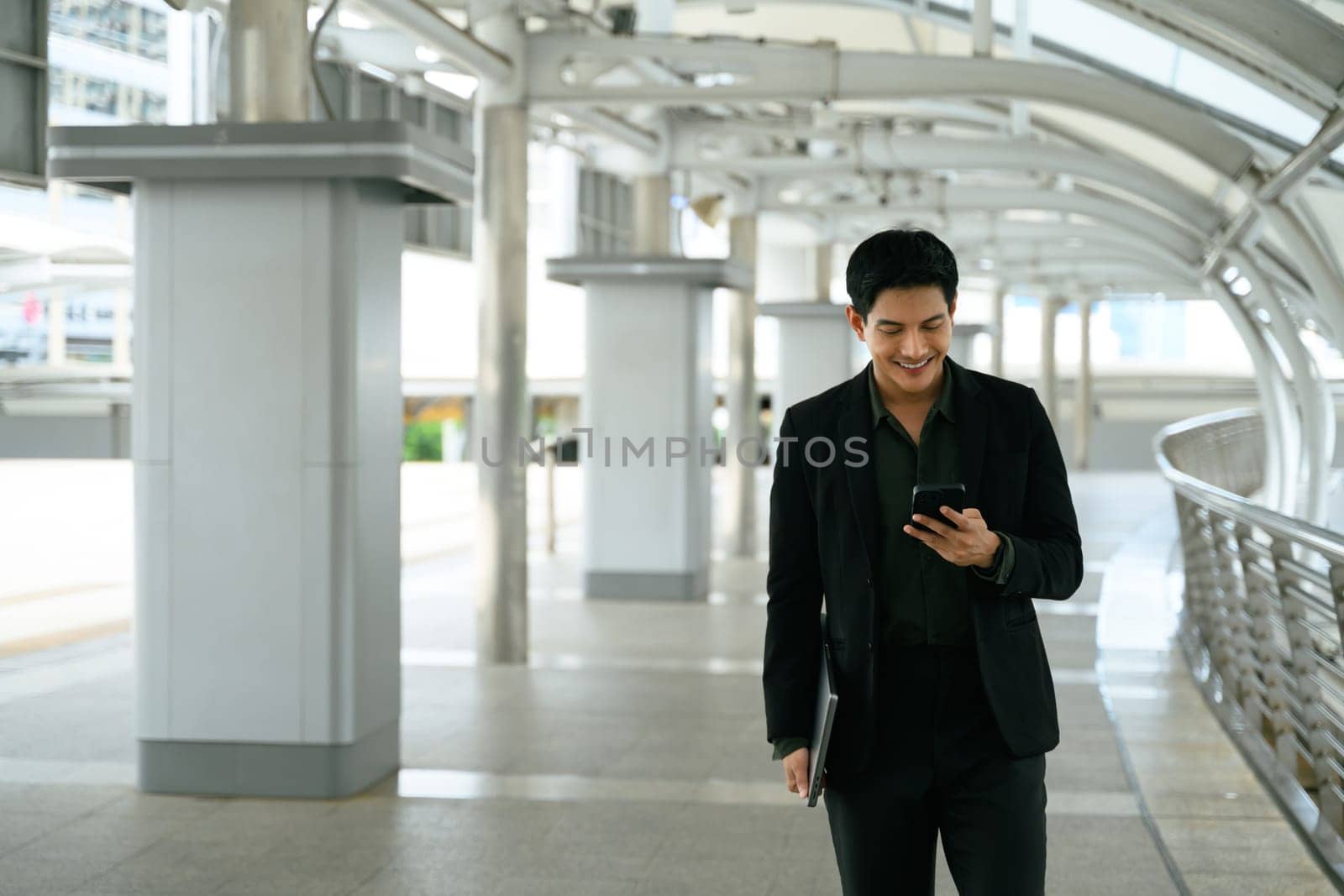 Confident executive walking through a modern city walkway and using smartphone.