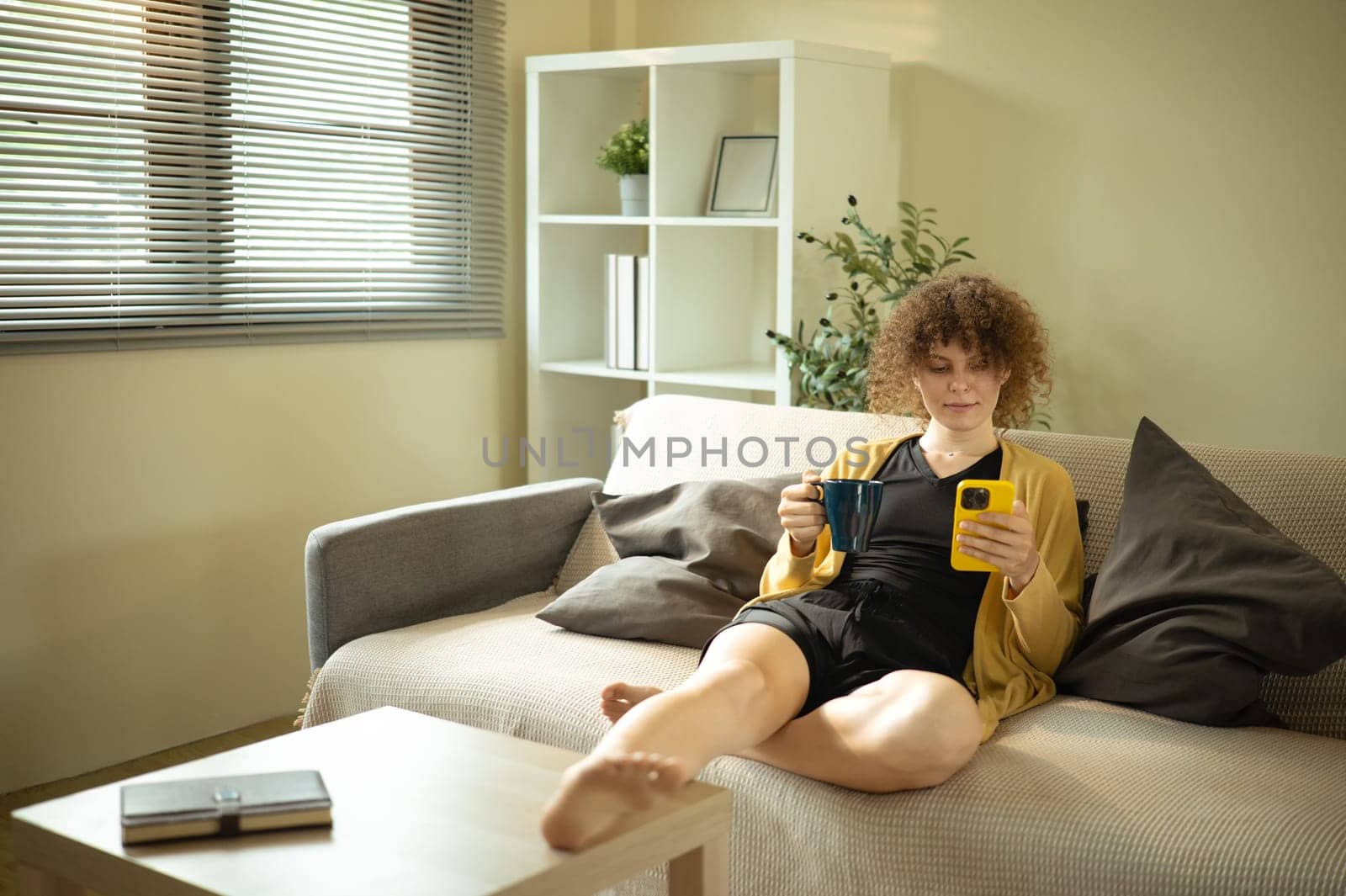 Relaxed caucasian woman sitting on couch at home holding a mug and checking her phone in living room.