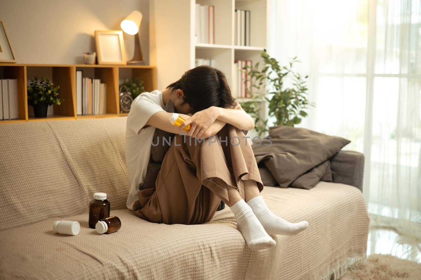 Depressed young man sitting on sofa, hugging knees surrounded by medicine bottles.
