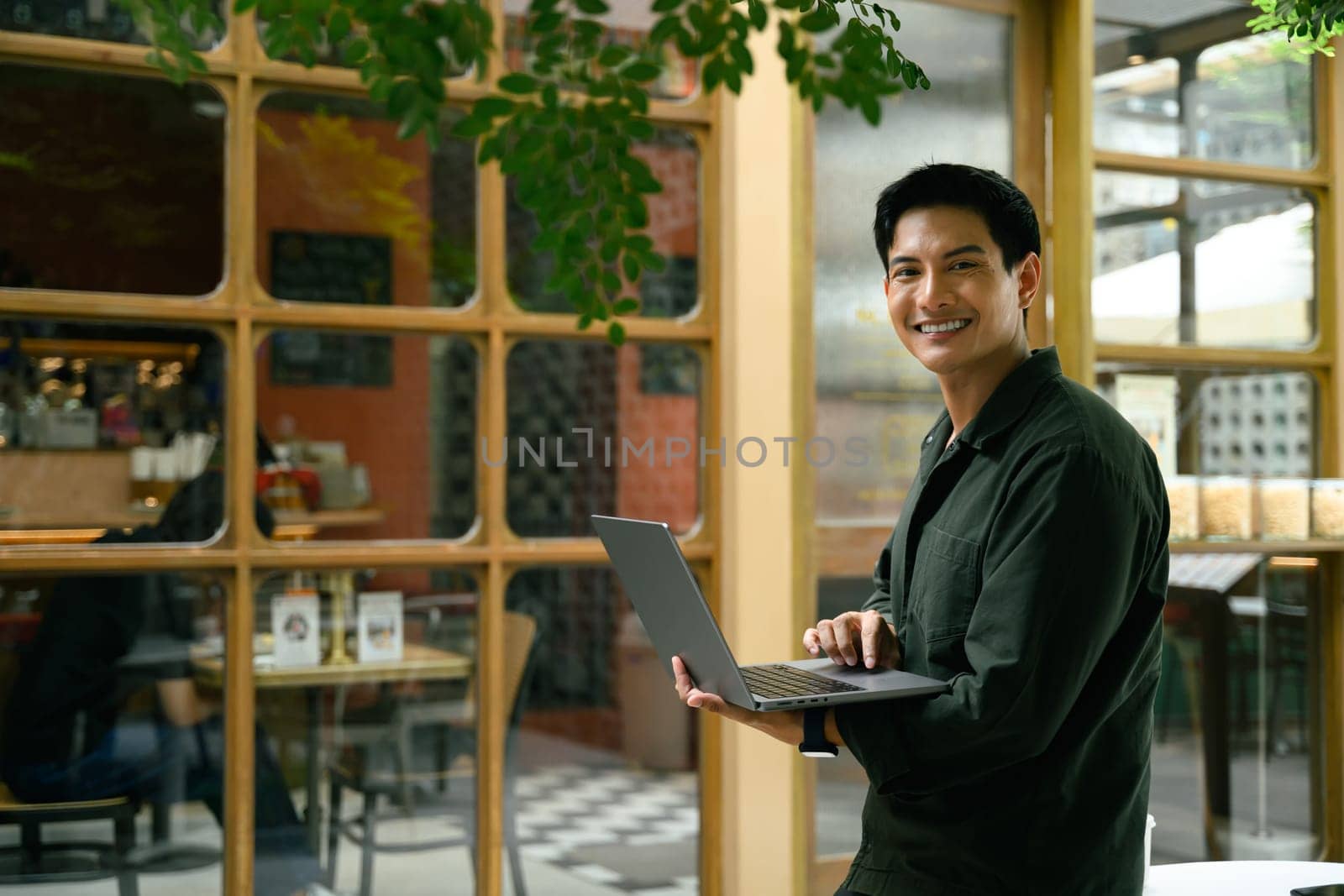 Smiling young man holding a laptop while standing outside an outdoor cafe.