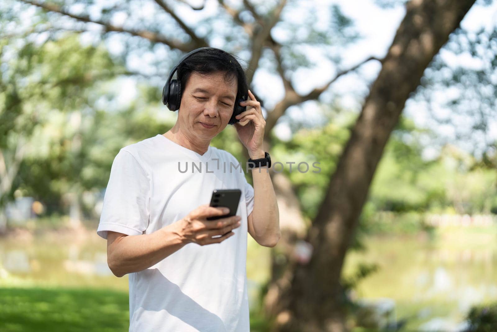An elderly man enjoys music on his smartphone, smiling in a serene outdoor environment, reflecting wellness and joy.