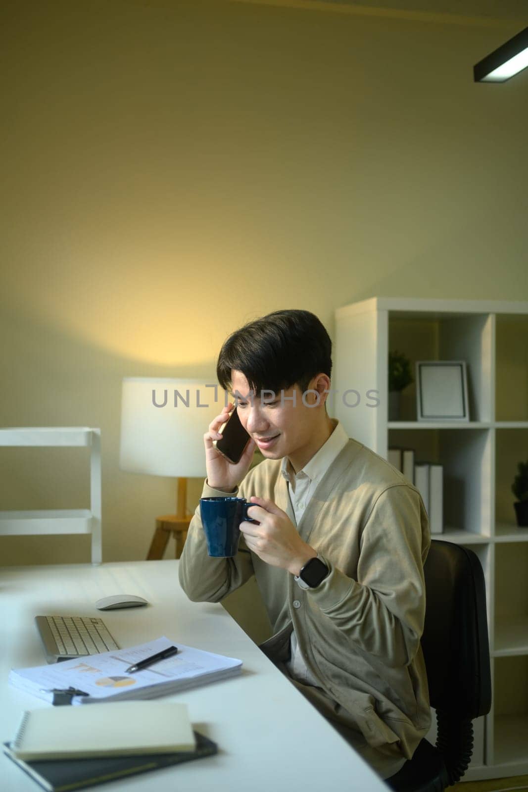 Young entrepreneur holding cup of coffee and talking on mobile phone at home office desk.
