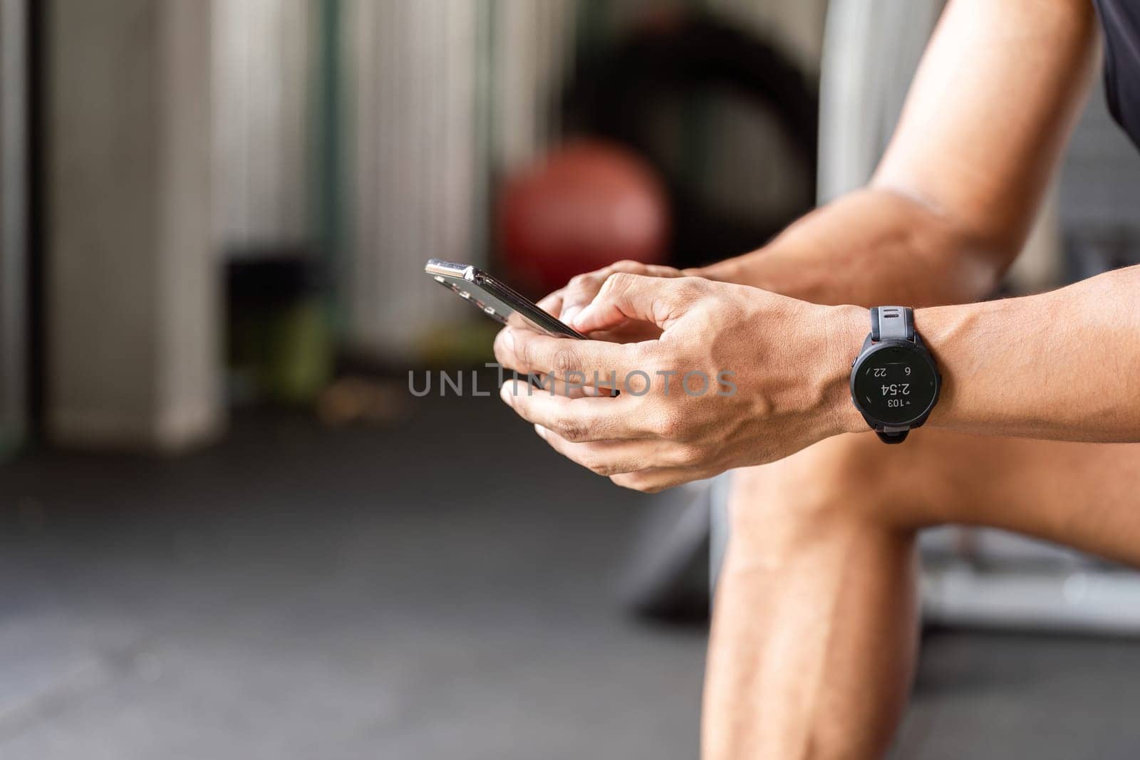 A man sits in the gym, focused on his smartphone, tracking his fitness progress and workouts using a fitness app.