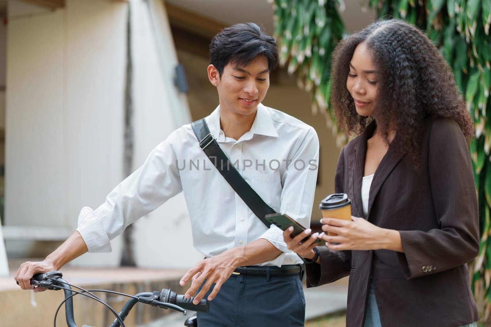 A young man and woman share insights on their digital devices, discussing sustainable transportation options, with a bicycle and coffee in a green outdoor setting.