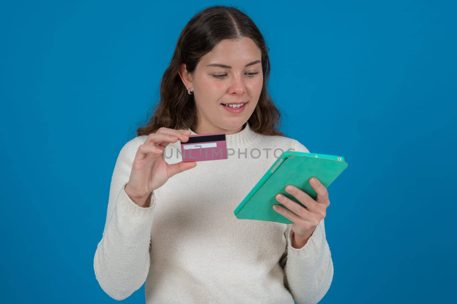 Young woman holding a credit card and a tablet, looking thoughtful.She wears a white sweater and stands against a blue background. Concept of online shopping, payment,or financial decisions.Copy space