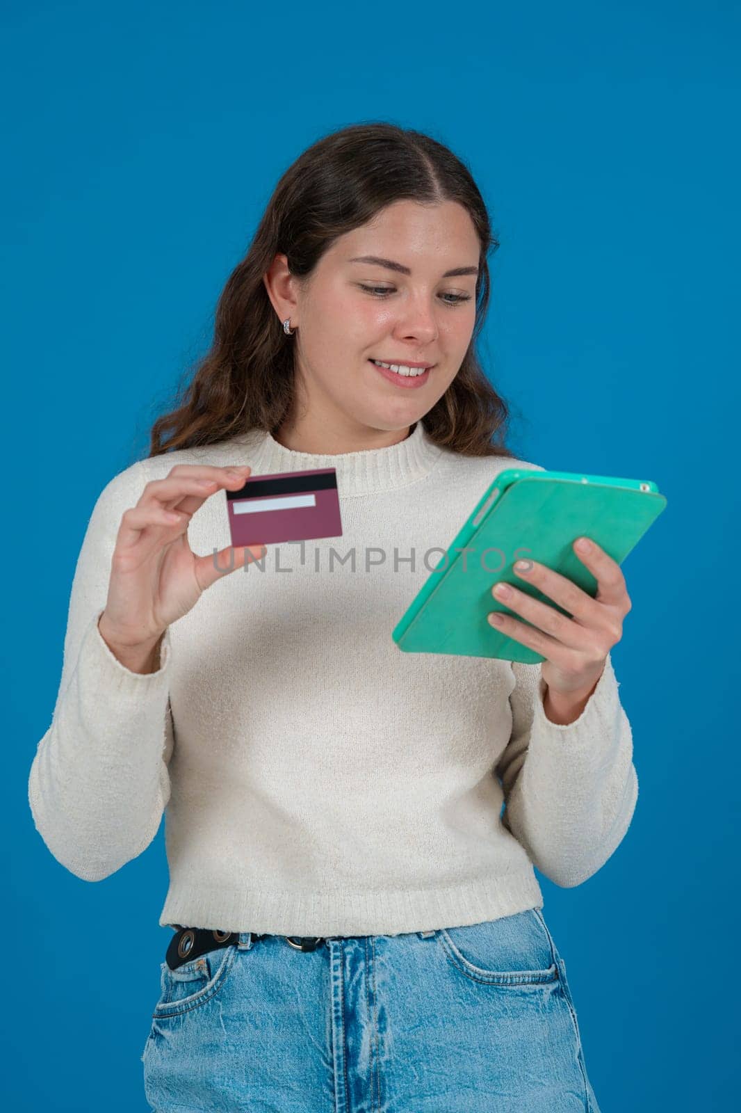 Young woman holding a credit card and a tablet, looking thoughtful.She wears a white sweater and stands against a blue background. Concept of online shopping, payment,or financial decisions.vertical