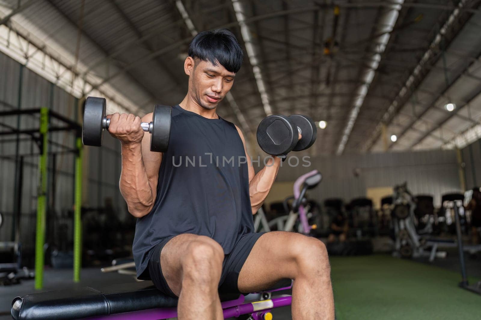 A focused young man lifts dumbbells for bicep curls in a gym setting, showcasing dedication to fitness and strength training.