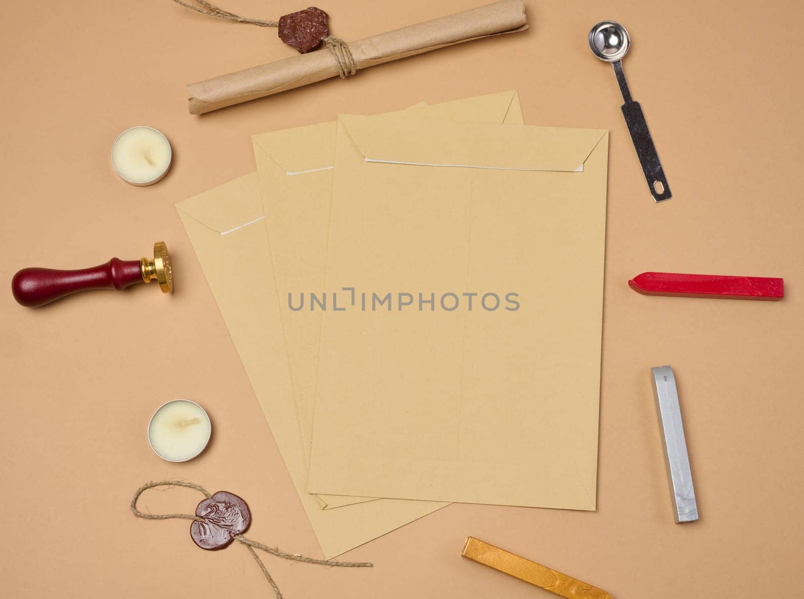 Wax seal kit including wax sticks, spoon, candles, stamp and brown envelopes placed on beige background, top view