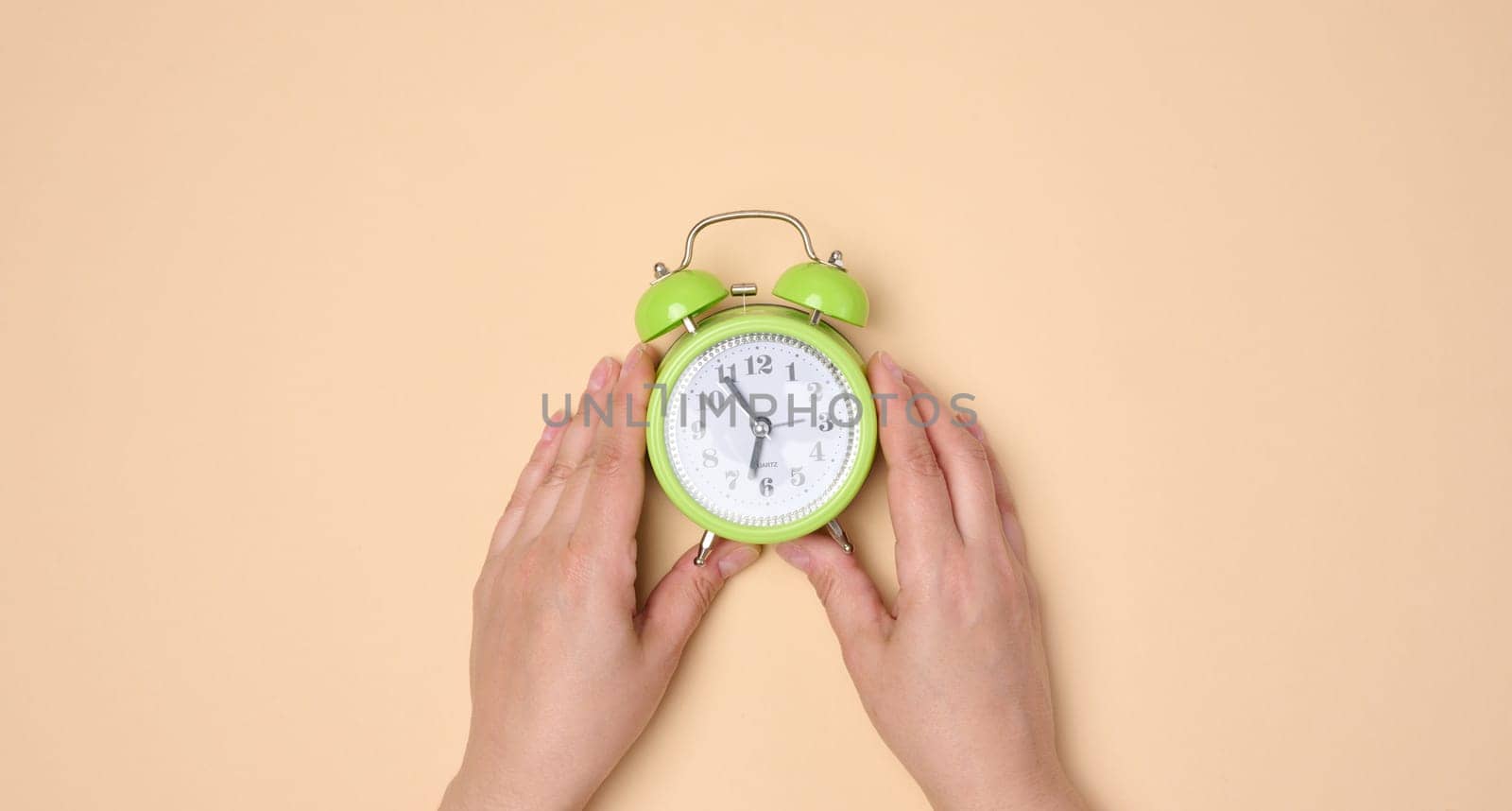 Hands holding green table alarm clock on neutral background.