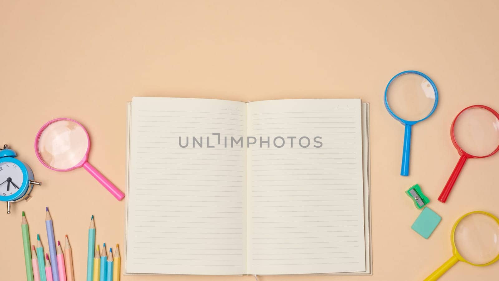 Top view of an open notebook with pastel-colored pencils, green sharpener, and turquoise eraser on a beige background. Flat lay composition of school or office stationery supplie