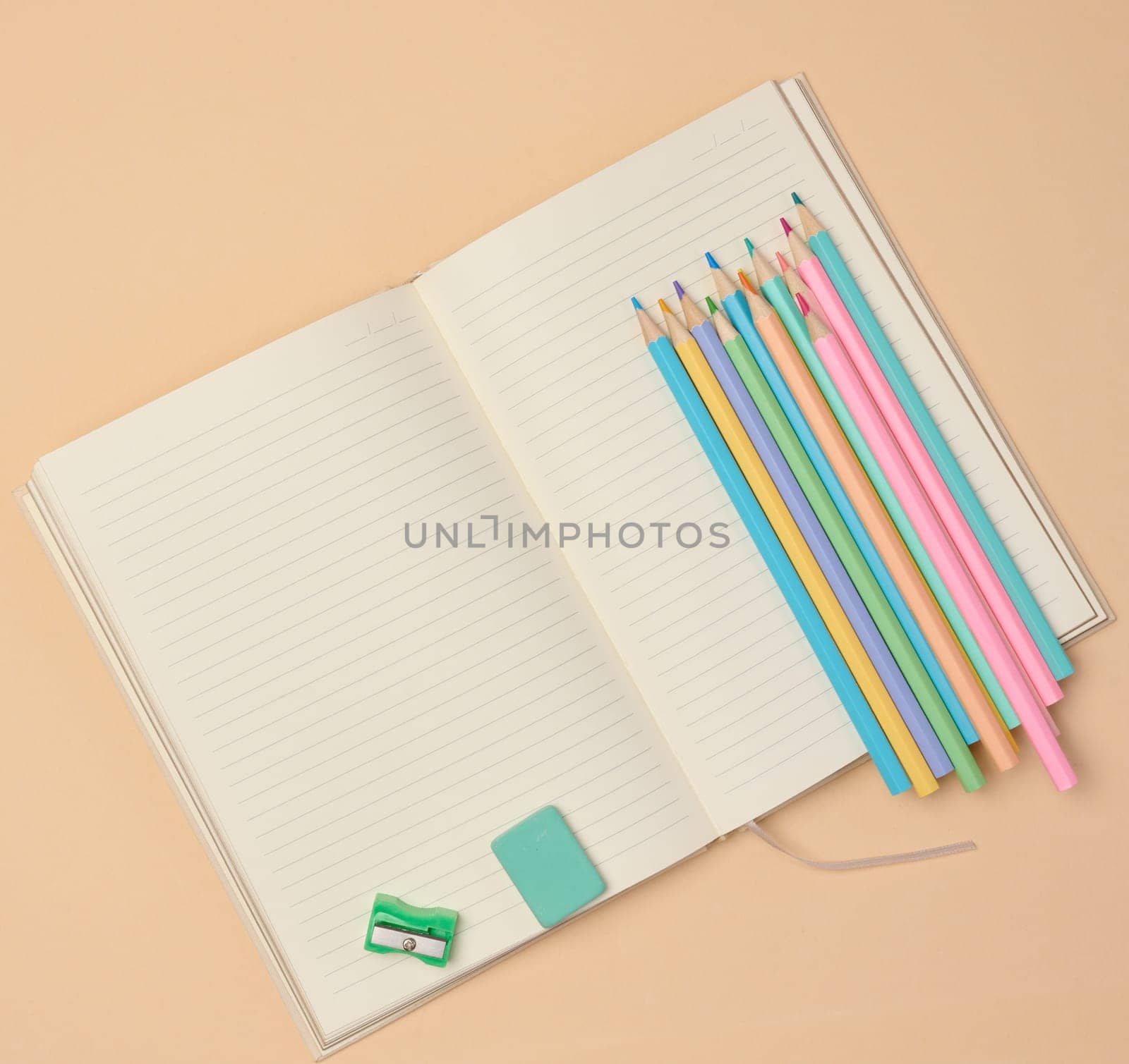 Top view of an open notebook with pastel-colored pencils, green sharpener, and turquoise eraser on a beige background. Flat lay composition of school or office stationery supplies.