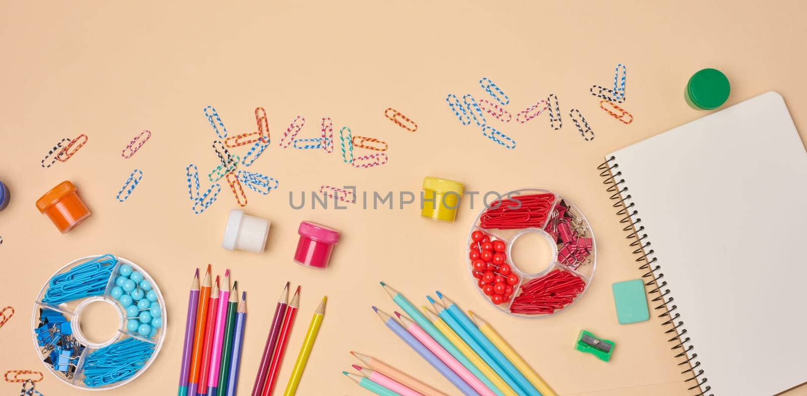 Flat lay of colorful school and office supplies including pencils, paper clips, notebooks, paints, and sharpeners on a peach background. Creative workspace concept.