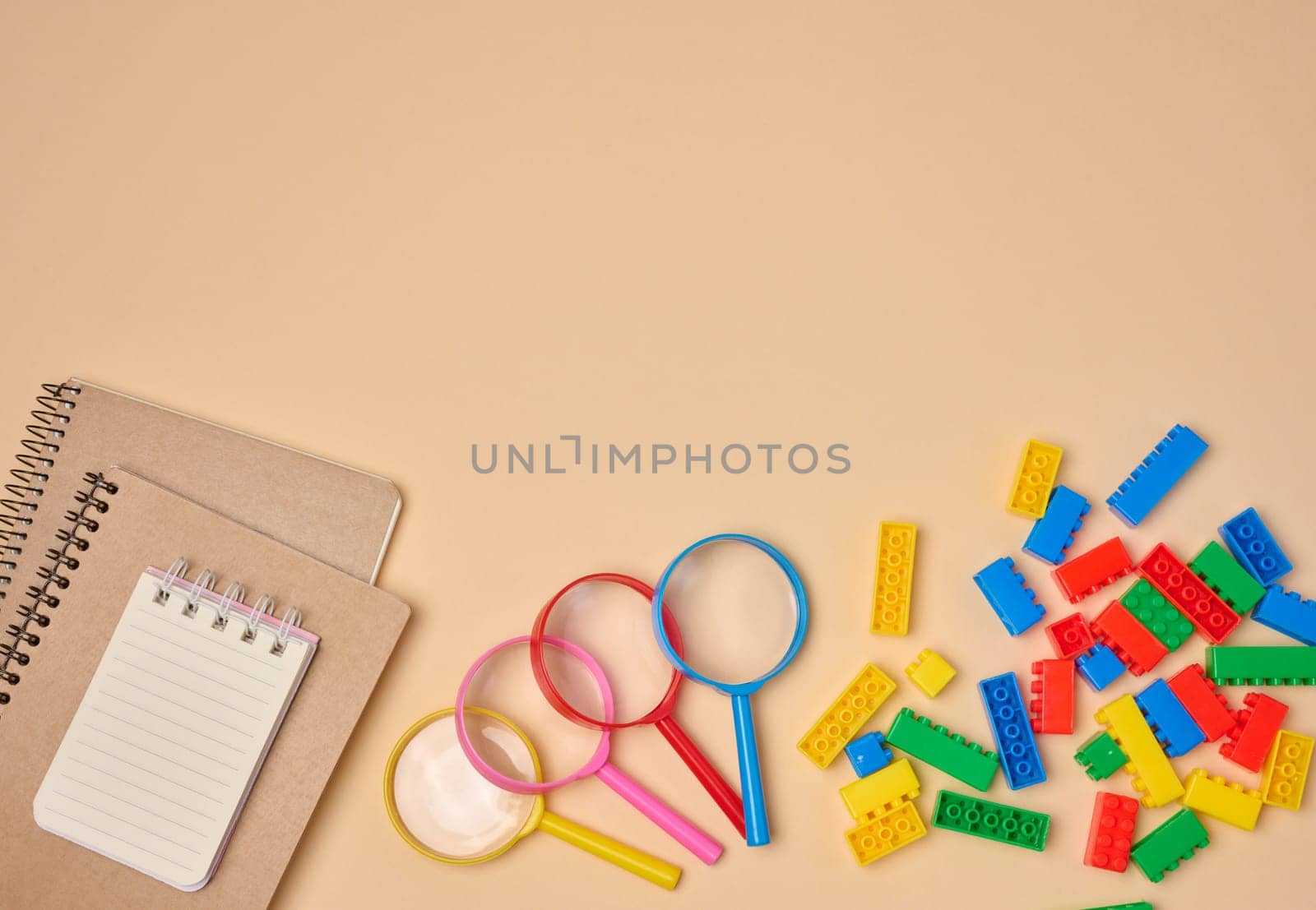 Flat lay of colorful plastic building blocks, magnifying glasses, and notebooks on a peach background. Educational and playful setup for children’s learning activities.