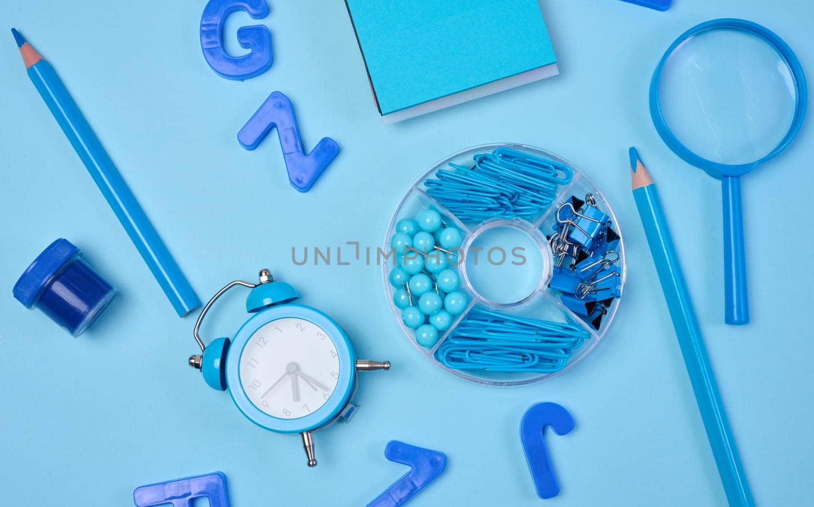 Flat lay of blue stationery, learning items letters, magnifying glass, supplies, and an alarm clock on a blue background. Education, time, and study concept