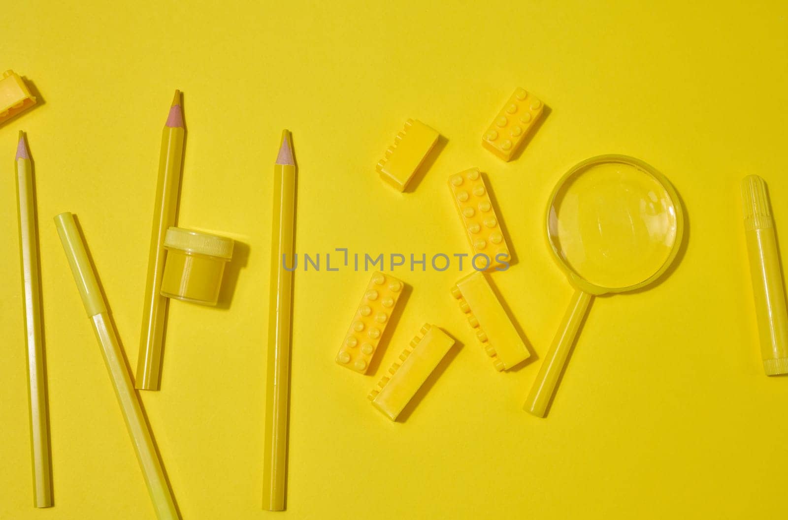 Collection of yellow stationery, building brick, and magnifying glass on a yellow background. Pencil, marker, jar included. Creative or learning concept.