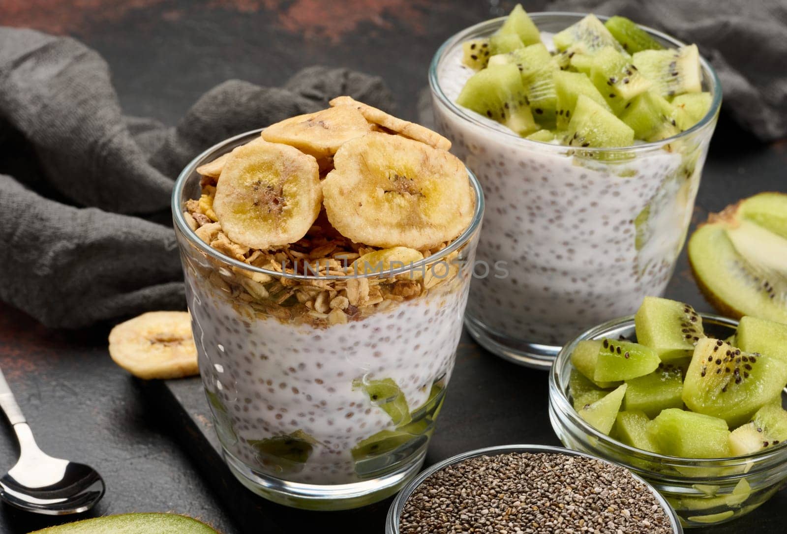 Two glasses of chia seed pudding, one topped with diced kiwi and the other with granola and banana chips, are on a dark surface with ingredients