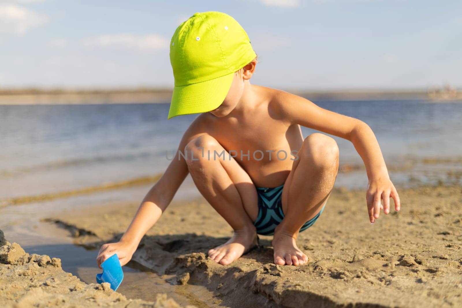 Child wearing bright cap and swimwear playing with a plastic scoop on a sandy beach near calm water on a sunny summer day.