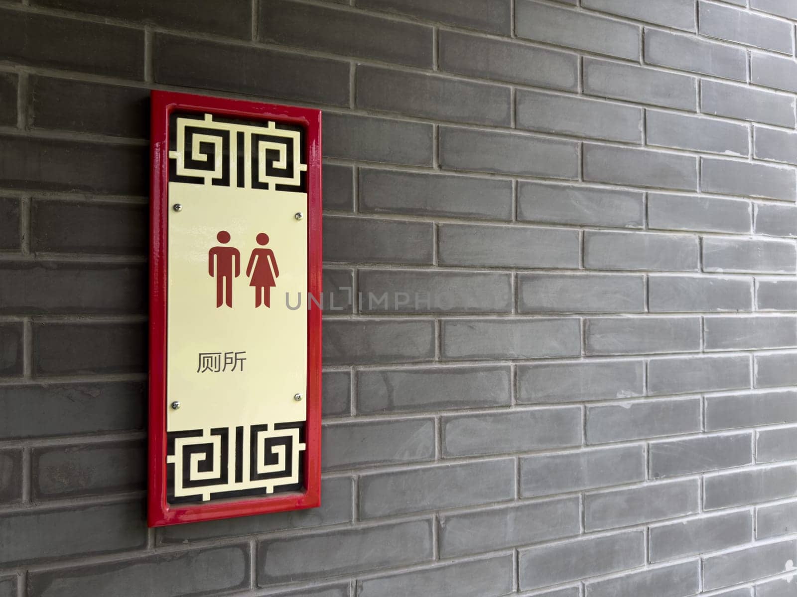 Restroom sign in Chinese style with traditional patterns and red accents, mounted on a gray brick wall. The sign includes male and female pictograms and Chinese characters indicating public toilet