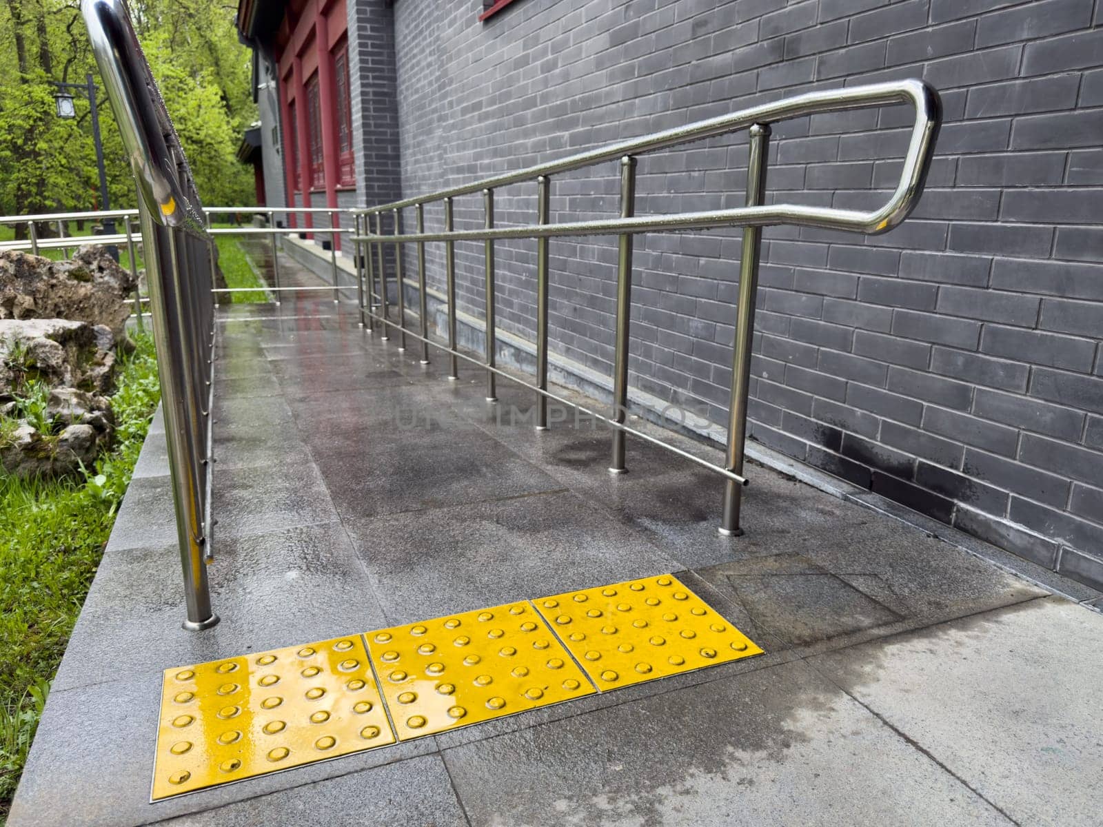 Tactile paving tiles with raised dots installed in front of accessibility ramp with metal handrails, providing guidance and safety for visually impaired individuals near building with brick wall