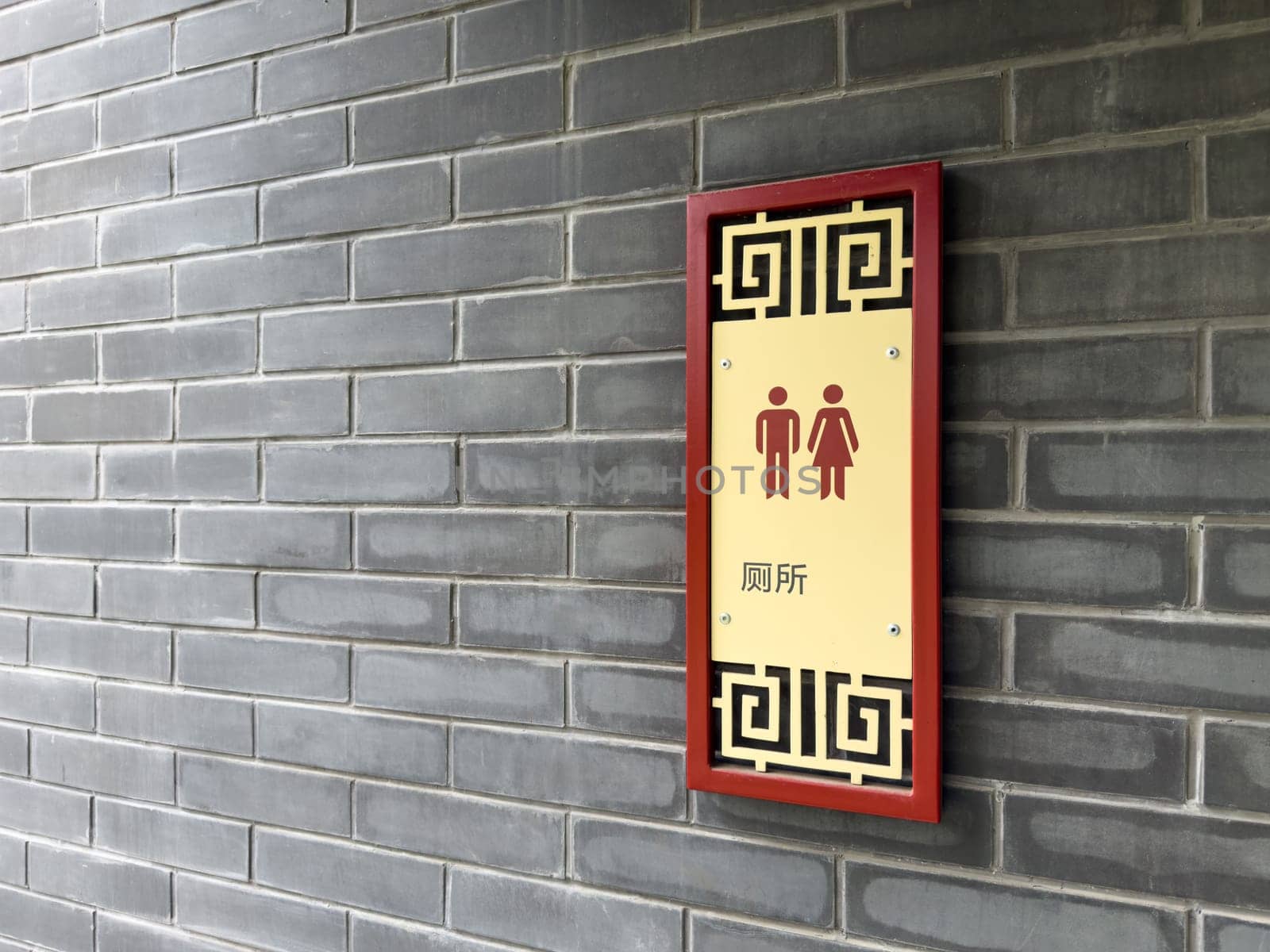 Wall mounted restroom sign with male and female pictograms and Chinese characters, framed with traditional Chinese ornament, attached to gray brick wall in an Asian inspired architectural setting