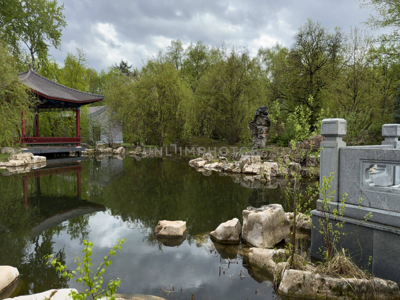 Scenic view of traditional Chinese garden with red pavilion by the pond, stone bridge elements, ornamental rocks and lush greenery reflecting in the still water. Serene and timeless scene