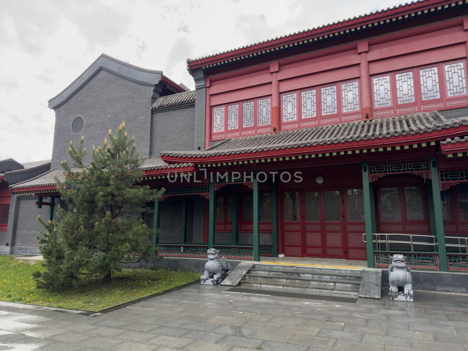 Traditional Chinese style building with red facade, tiled roof, decorative lattice windows and stone guardian lion statues at the entrance. Classic architectural elements dominate the composition