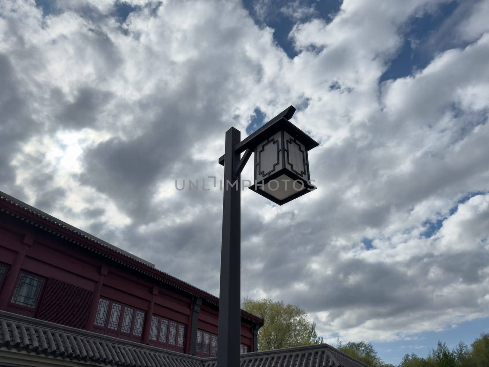 Traditional Chinese style lantern on a tall pole against dramatic cloudy sky and tiled roof. Minimalist architectural detail emphasizing cultural design and outdoor ambiance