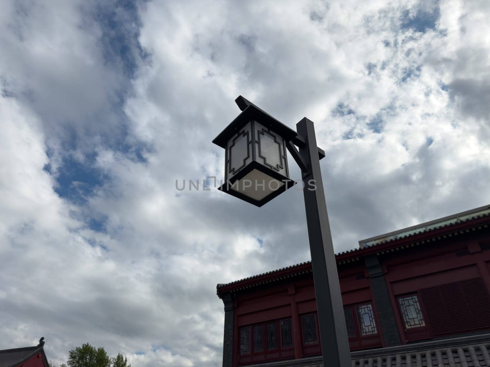 Traditional black and white lantern on a tall post stands in front of red Chinese style building with decorative windows and cloudy sky, emphasizing cultural design and symmetry