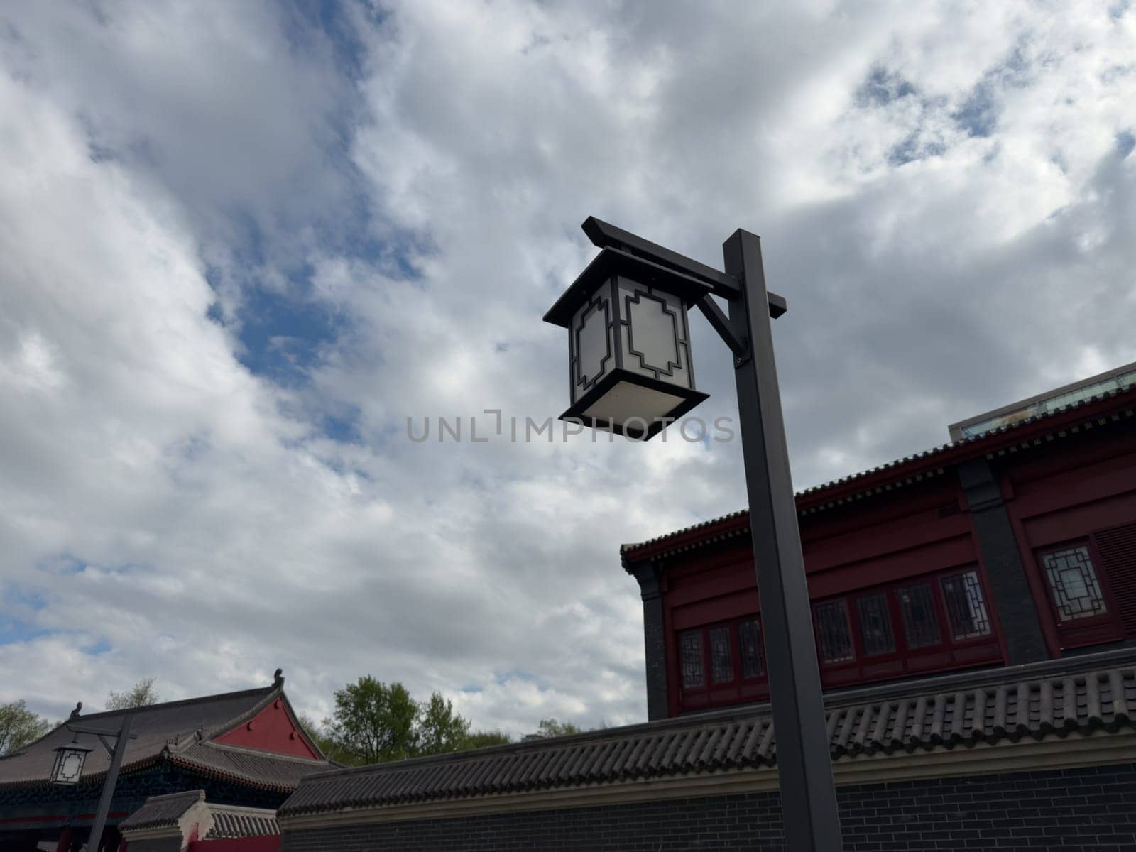 Modern street lantern with traditional Chinese design elements stands near red roofed architecture under a cloudy sky, blending cultural heritage with functional urban lighting