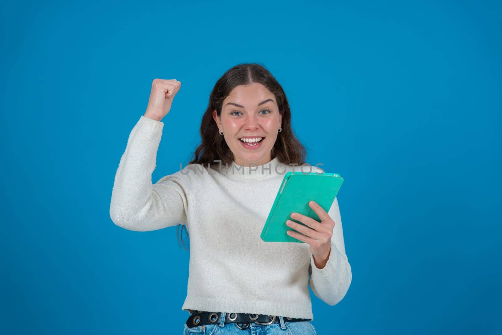 Happy young woman in a white sweater raising her fist in celebration while holding a green tablet and smiling at the camera, against a blue background, symbolizing success, joy, and digital life.