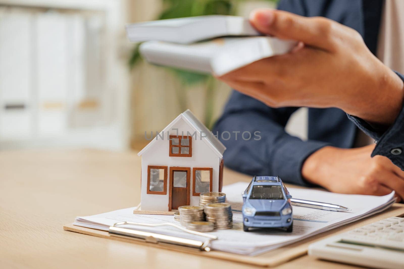 Closeup of hands holding bills near a car and house model. Concept of cash savings, property tax planning, banking, and loans to achieve financial goals for purchasing assets like homes and vehicles.