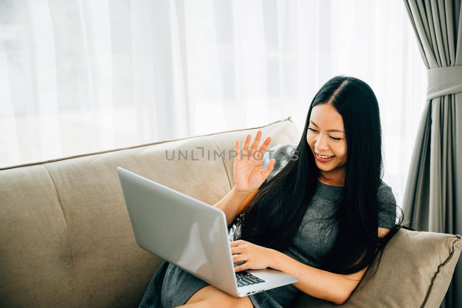 Woman on sofa uses laptop for video conferencing saying hi to friend. Engaged in distant meeting discussing laughing and listening. Modern online communication and connection.