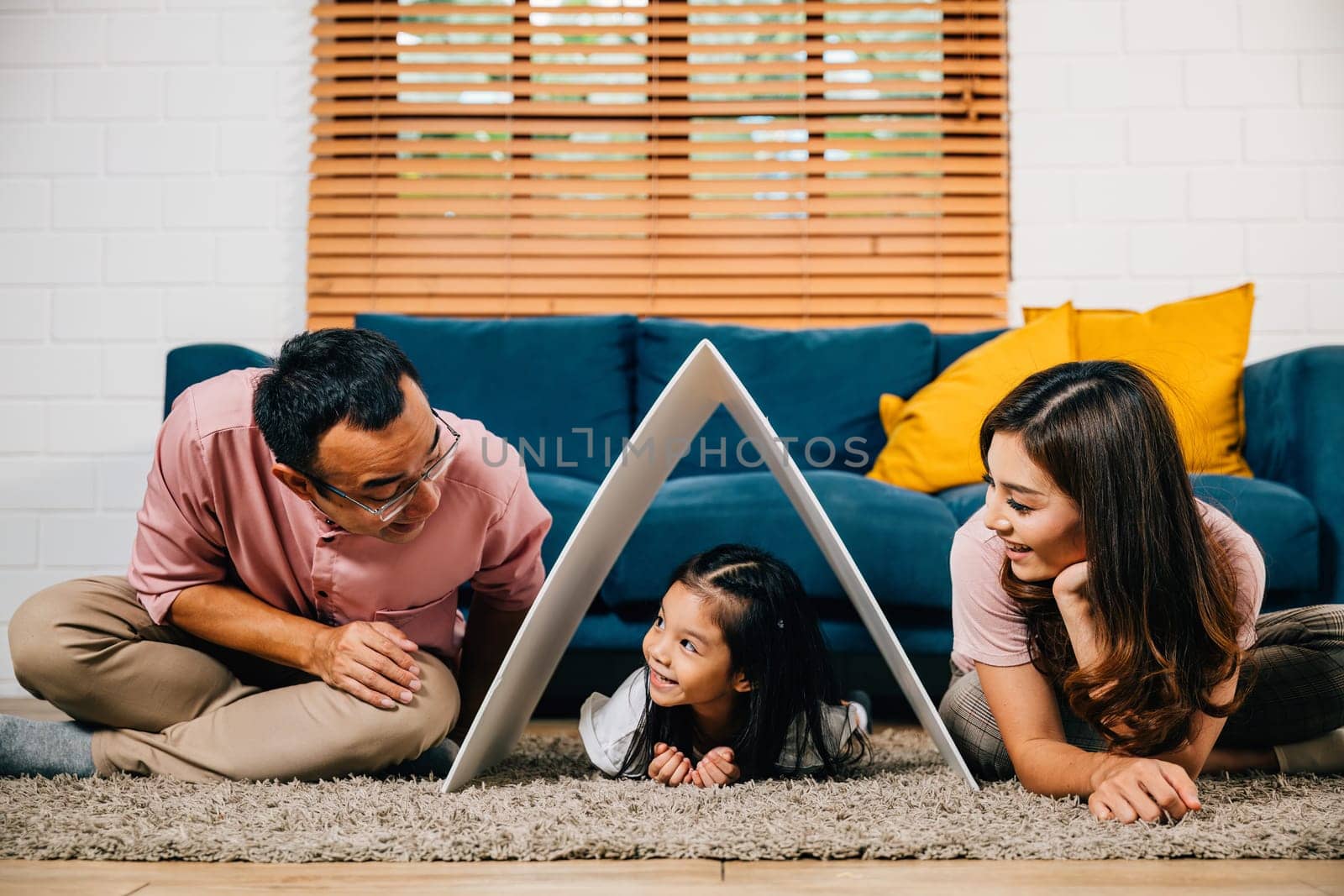 In their new home a cheerful family on a couch holds a cardboard roof symbolizing investment and happiness. Asian parents and their daughter showcase security and togetherness.