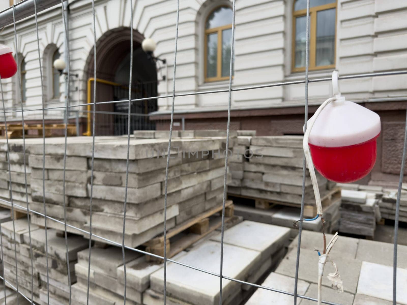 Urban construction scene with stacked paving slabs behind a metal fence and a small red warning light. The image represents infrastructure upgrades and street surface renovation work